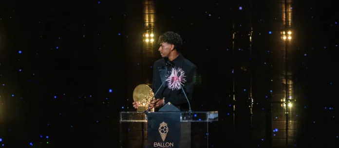 Seorang pesepak bola muda mengenakan jas hitam memegang trofi emas di podium Ballon d'Or