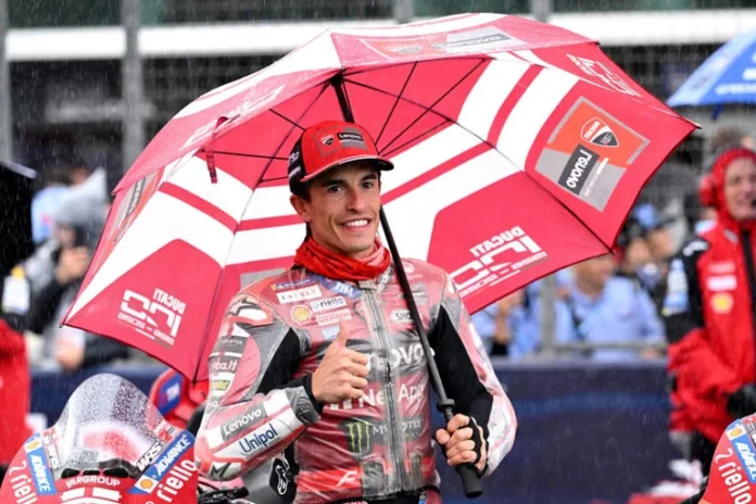 Pembalap Ducati mengenakan perlengkapan balap merah memegang payung di area sirkuit dan mengacungkan jempol